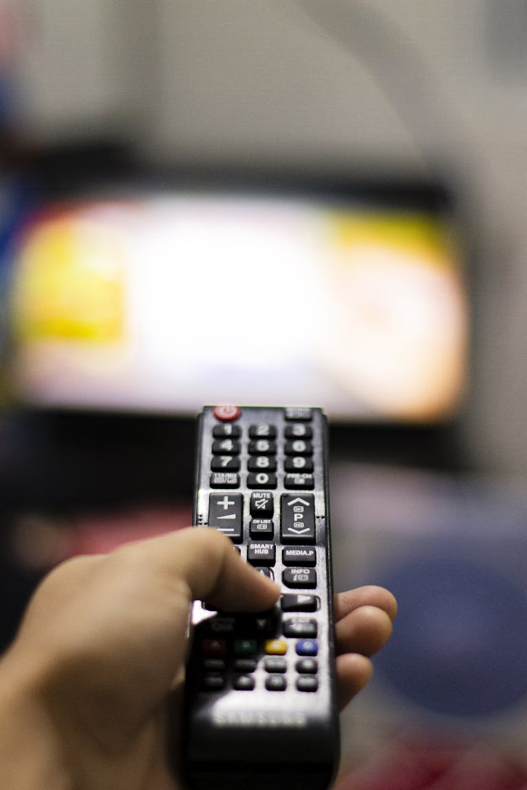 Close-up of a hand holding a remote control, watching TV indoors. Captures modern leisure and technology.